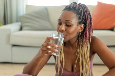 Domestic sports and hydration. Young african american woman drinking fresh water from glass, resting after exercising at home in living room, free space
