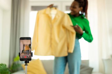 Fashion blog. African american female influencer recording video content at home, making review of new clothes in front of camera, selective focus on cellphone
