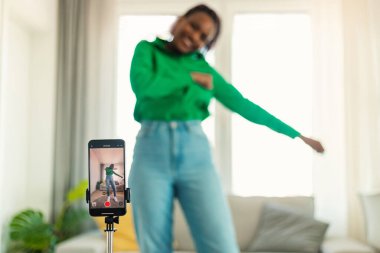 Young black woman recording video content, dancing on camera at home, selective focus of smartphone on tripod, free space. Positive lifestyle vlogger filming for social media