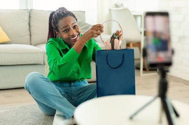 Popular black female blogger shooting video review of new headphones, talking to cellphone camera on tripod at home. Woman recommending modern gadget, broadcasting for social network