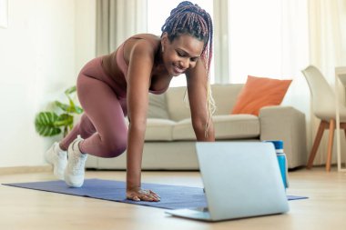 Training at home. Fit black lady doing cross body mountain climbers exercise, watching online tutorial on laptop. Woman exercising in living room at home