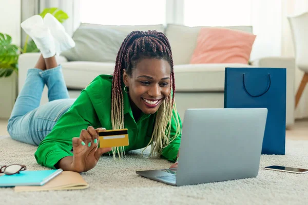 Its time for shopping. Happy african american woman using laptop and holding credit card, making purchases, buying gifts and holiday presents, lying on the floor carpet