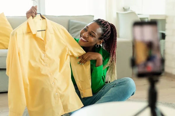 Happy black lady recording video review of new clothes, talking to smartphone camera in living room interior. Fashion blog, online shopping, sale, emotions concept