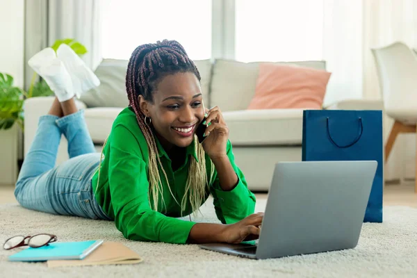 Positive black woman with gift bags shopping online on laptop, confirming web order on smartphone from home, lying on floor carpet. Happy lady buying goods on internet