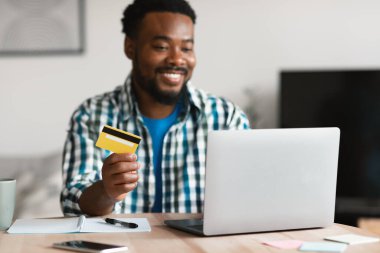 Smiling Black Man Shopping Online At Laptop Computer Holding Credit Card Sitting At Desk At Home. Guy Making Payment Via Internet Banking Service. Ecommerce And Finances Concept. Selective Focus