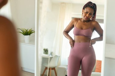 Unhappy black woman looking at her reflection in mirror and feeling upset with results of slimming diet, not losing weight, disappointed about her body parameters at home