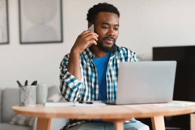 African American Freelancer Guy Calling By Cellphone Working Online On Laptop Communicating Sitting At Home. Business Communication And Entrepreneurship Concept. Selective Focus