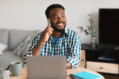 Cheerful Black Guy Talking On Phone Using Laptop Working Online Sitting At Home, Looking Aside. Male Freelancer Communicating By Cellphone. Freelance And Business Communication