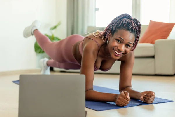Body shaping workout. Fit black woman standing in plank pose with leg lift, exercising on yoga mat and watching video tutorial on laptop, smiling at camera