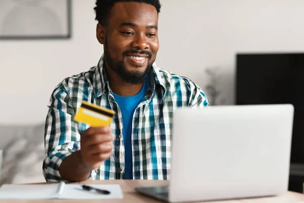 Online Shopping. Happy Black Buyer Man At Laptop Holding Credit Card Making Payment Via Internet Banking Service At Home. Retail And Ecommerce Concept. Selective Focus