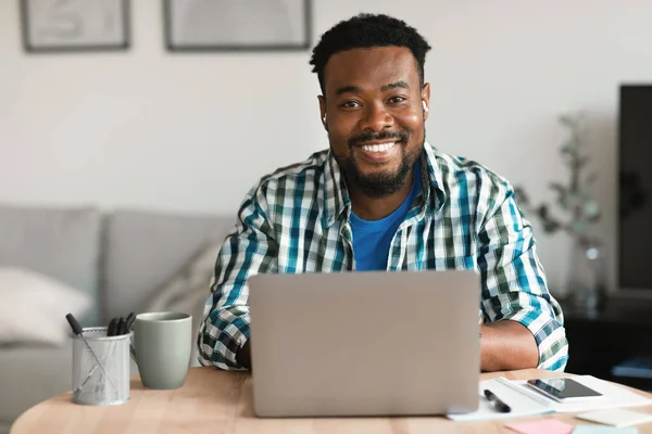 Cheerful Black Man At Laptop Computer Posing Wearing Earbuds Working Online Sitting At Desk Smiling To Camera Indoors. Entrepreneurship Career And Internet Technology Concept