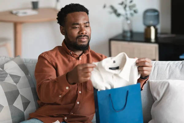 Discontented African American Male Buyer Unpacking Shopper Bag With New Clothes After Unsuccessful Shopping Sitting On Sofa At Home. Male Fashion And Ecommerce Problem. Selective Focus