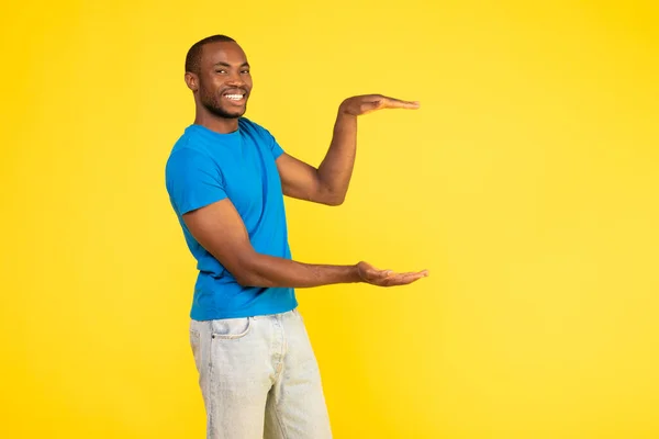 Cheerful African American Male Holding Invisible Object In Hands Advertising New Product Smiling To Camera Standing Over Yellow Background In Studio. Look At This