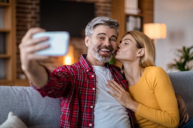 Smiling middle aged caucasian wife kisses husband on cheek, man with beard make selfie on smartphone in room interior. Photo for social networks and blog, video call, love and relationship at home