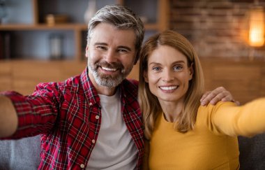 Cheerful middle aged caucasian male and female look at camera, have fun, make photo in living room interior. Video call, selfie for social networks and blog, love, relationship romance and family