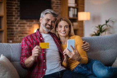 Smiling adult caucasian man hug lady with smartphone and credit card, sitting on sofa, shopping online in living room interior. Ad and online offer, technology for finance, sale for shopaholic at home