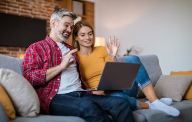 Smiling mature caucasian couple waving hand in laptop, have meeting together, sitting on sofa, in living room interior. New normal at home, video call and social distance with gadget due covid-19