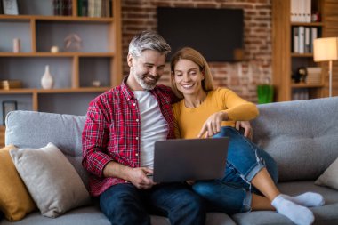 Cheerful mature caucasian wife show to man to laptop, watching video together, sit on sofa, in living room interior. Sale for shopaholic at home, ad and internet offer, video call and modern gadget