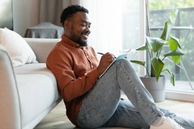 Cheerful Black Male Writer Writing And Taking Notes In Notebook Sitting On Floor At Home On Weekend. Creative Writing And Hobby Concept. Side View Shot
