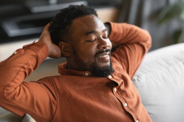 African American Man Sitting Holding Hands Behind Head Relaxing With Eyes Closed At Home. Male Resting On Sofa On Weekend. Comfort And Relaxation Leisure Concept. Selective Focus