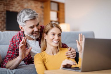 Glad smiling middle aged caucasian husband embracing wife, waving hand in laptop, making online call in living room interior. Communication remotely, video meeting at home during covid-19 quarantine