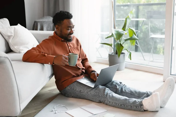 Black Freelancer Man Using Laptop Working Distantly Drinking Coffee Sitting On Floor At Home. Millennial Male Browsing Internet On Computer Indoor. Side View Shot