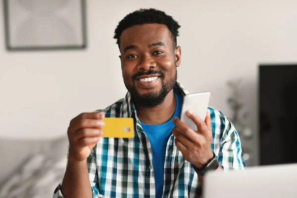 Cheerful Black Male Buyer Shopping Via Phone And Credit Card Online Sitting Smiling To Camera At Home. Guy Making Payment Via Bank Application. Selective Focus