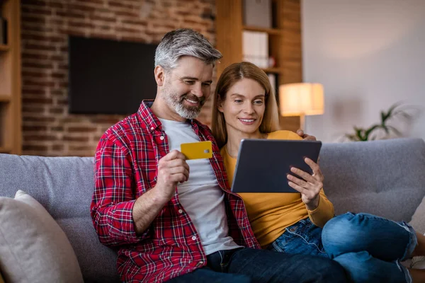 Cheerful mature caucasian husband and wife with tablet and credit card sitting on sofa shopping online in living room interior. Sale for shopaholic at home, ad and online offer, technology for finance