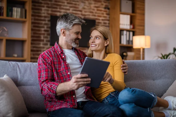 Smiling middle aged caucasian husband and wife with tablet sit on sofa, have video call in living room interior. Meeting remotely, ad and online offer, technology for communication and new normal