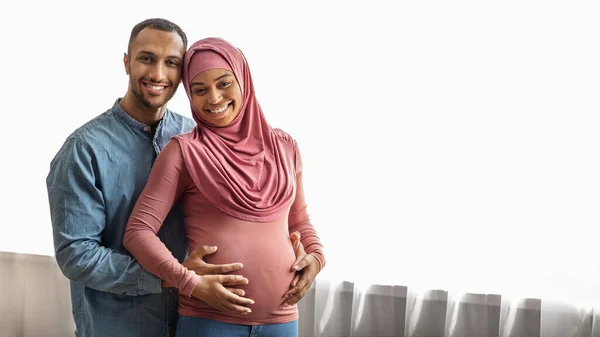 Pregnancy Concept. Portrait Of Romantic Black Islamic Couple Expecting Baby Embracing And Smiling At Camera While Posing Near Window At Home, Happy Muslim Spouses Enjoying Upcoming Parenthood