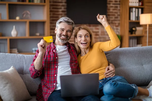 Happy adult caucasian couple with laptop, credit card rejoice to victory, successful purchase, sit on sofa, shopping online in room interior. Sale at home, ad and internet offer, win celebration