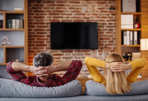 Smiling middle aged caucasian husband and wife relax, watch tv with blank screen and enjoy free time together in living room interior. Couple watch movie, show or match. Entertainment at home, back