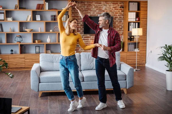Smiling middle aged caucasian husband and wife dancing, have fun and enjoy free time together in living room interior. Couple celebrating holiday, anniversary at home during covid-19 quarantine