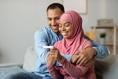 Happy black muslim couple checking pregnancy test while sitting on couch at home, cheerful young islamic spouses celebrating positive result, getting ready for upcoming parenthood, free space