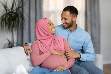 Loving pregnant black islamic spouses bonding together at home, happy awaiting muslim family embracing while relaxing on couch in living room, hugging and smiling to each other, free space
