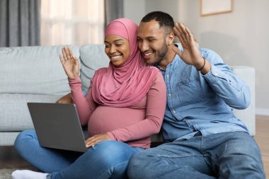 Black Muslim Spouses Expecting Baby Making Video Call With Laptop At Home, Happy Pregnant Islamic Couple Teleconferencing With Friends Or Family Via Computer, Waving Hand At Web Camera,