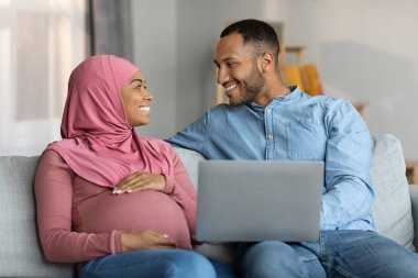 Pregnant Black Muslim Couple Resting With Laptop Computer At Home, Happy Islamic Spouses Sitting On Sofa With Modern Gadget And Smiling, Using Internet While Relaxing Together In Living Room