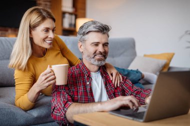 Happy mature caucasian wife and husband looking at laptop, enjoy coffee break in free time in living room interior. Video call, meeting online, modern technology, social network and blog at home