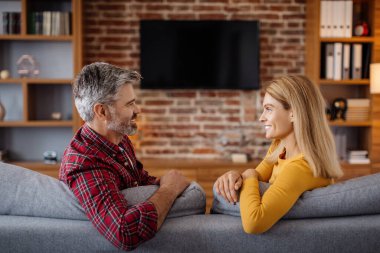 Cheerful mature caucasian female and male relaxing, talking, watching tv with empty screen and enjoying free time together in living room interior. Couple watch movie, show or ad at home together