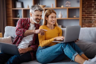 Happy mature caucasian man look at wife with laptop, chatting, watch video, ad and offer in living room interior. Work and study at home, gadget in free time, technology during covid-19 quarantine