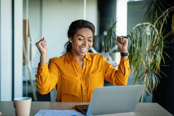 Business success. Overjoyed black woman using laptop and gesturing yes, shaking fists celebrating big luck, sitting at workplace in modern office