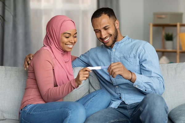 Parenthood Concept. Happy Black Muslim Spouses Holding Positive Pregnancy Test While Sitting On Couch At Home, Smiling Islamic Couple Getting Ready For Childbirth, Celebrating Good News