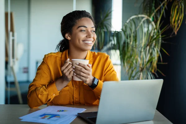 Dreamy Black Businesswoman Using Laptop Holding Coffee Cup Looking Aside — Stockfoto