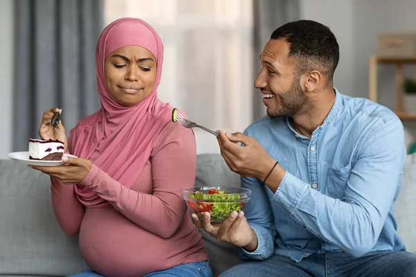 Caring Black Man Offering Fresh Vegetable Salad To His Pregnant Muslim Wife Eating Cake While They Sitting Together On Couch At Home, Islamic Lady Frowning, Choosing Dessert Over Healthy Nutrition