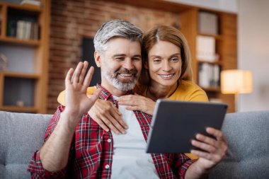 Cheerful mature caucasian female hugging man, waving hand, watching video in tablet, calling online in living room interior, close up. New app for remote communication, meeting at home during covid-19