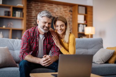 Cheerful middle aged caucasian man and woman watch video in laptop, making online call in living room interior. Communication remotely, meeting at home, ad and online offer during covid-19 pandemic
