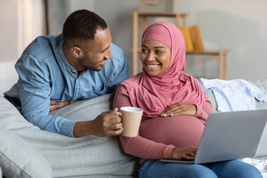 Portrait Of Happy Islamic Couple Expecting Baby Relaxing With Laptop At Home, Cheerful Black Muslim Spouses Using Computer While Resting Together In Living Room Interior, Enjoying Weekend Leisure