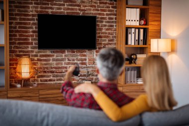 Adult caucasian man with remote control and lady relaxing, watching tv with blank screen and enjoy free time in living room interior. Couple and movie night at home together, entertainment with device