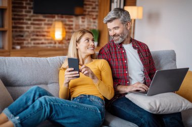 Glad adult caucasian female show phone to man with laptop, watch video in living room interior. New app, work and study at home, rest with gadget in free time at home, great offer and ad due covid-19