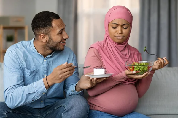 Husband Offering Cake To Pregnant Muslim Wife, Woman Frowning And Eating Salad, Expecting Islamic Woman In Hijab Choosing Healthy Nutrition During Pregnancy, Preferring Vegetables Over Sweets